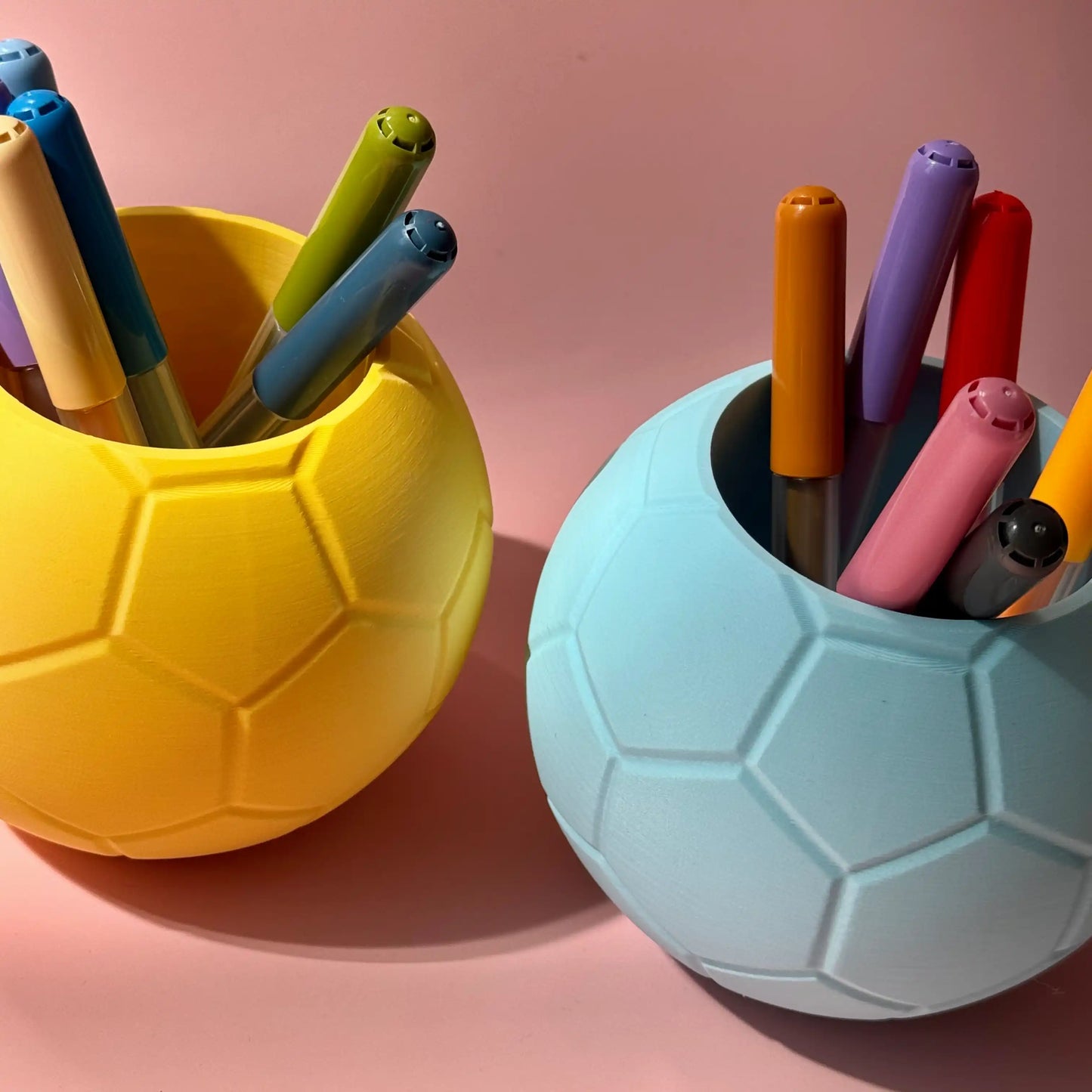Stiftebox Fussball in hellblau und gelb gefüllt mit Stiften vor rosa Hintergrund